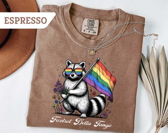 May include: A brown t-shirt with a graphic of a raccoon wearing rainbow sunglasses and holding a rainbow pride flag. The text "Fox Trot Delta Tango" is printed below the raccoon.