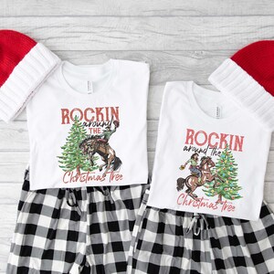 May include: Two white t-shirts with a Christmas tree design and the text "Rockin' around the Christmas Tree". The design features a person riding a horse around a decorated Christmas tree. The shirts are paired with black and white plaid pajama pants and red and white Santa hats.