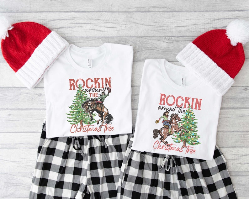 May include: Two white t-shirts with a Christmas tree design and the text "Rockin' around the Christmas Tree". The design features a person riding a horse around a decorated Christmas tree. The shirts are paired with black and white plaid pajama pants and red and white Santa hats.