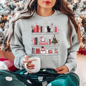 May include: A grey sweatshirt with a Christmas-themed design featuring a bookshelf with books, decorations, a snowman, Santa Claus, and a cat. The design is in a cartoon style.