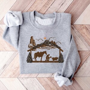 Puede incluir: Sudadera gris claro con un gr&aacute;fico marr&oacute;n de un pesebre con un caballo, monta&ntilde;as y una estrella. El dise&ntilde;o tiene un tema r&uacute;stico y occidental, con un estilo de silueta. La sudadera est&aacute; hecha de un material suave.