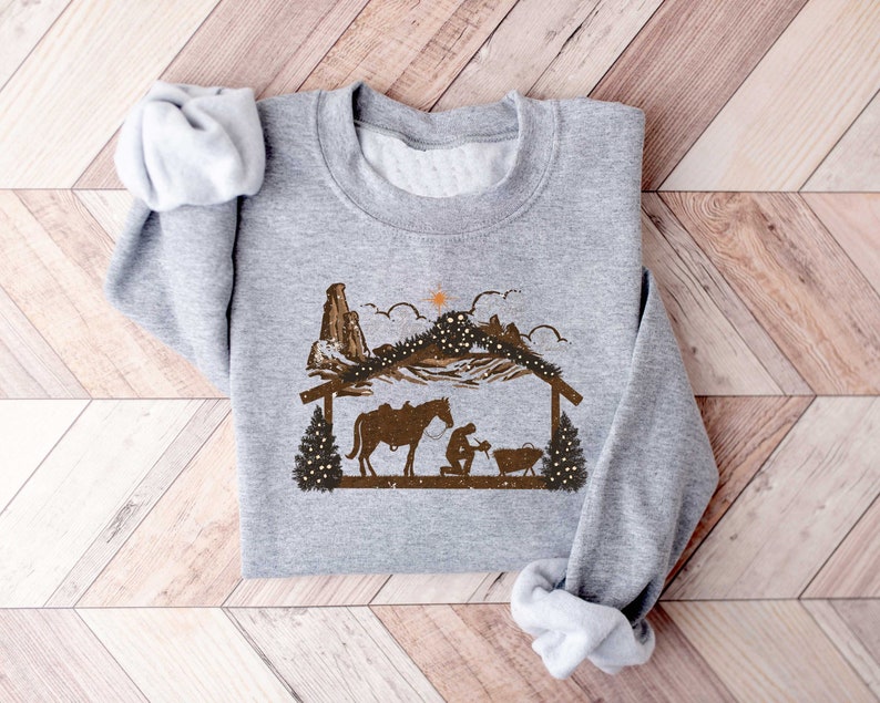 Puede incluir: Sudadera gris claro con un gr&aacute;fico marr&oacute;n de un pesebre con un caballo, monta&ntilde;as y una estrella. El dise&ntilde;o tiene un tema r&uacute;stico y occidental, con un estilo de silueta. La sudadera est&aacute; hecha de un material suave.