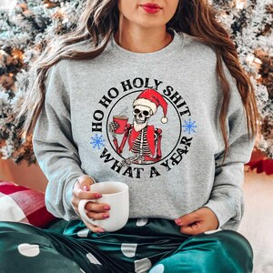 May include: A gray sweatshirt with a white circle graphic featuring a skeleton wearing a Santa hat and holding a mug. The text inside the circle reads "Ho Ho Holy Shit What A Year".