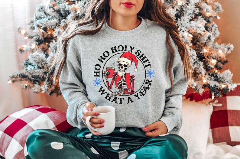 May include: A gray sweatshirt with a white circle graphic featuring a skeleton wearing a Santa hat and holding a mug. The text inside the circle reads "Ho Ho Holy Shit What A Year".