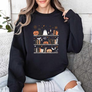 May include: A black sweatshirt with a graphic of a bookshelf filled with books, pumpkins, a black cat, a ghost, and candles. The graphic is in a vintage style with a white background and black outlines.