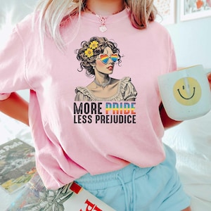 May include: Pink t-shirt with the text "MORE PRIDE LESS PREJUDICE" and a graphic of a woman wearing rainbow heart-shaped sunglasses. The woman has yellow flowers in her hair. The shirt is a light pink color.