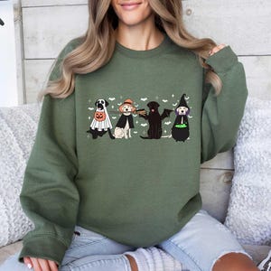 May include: A green sweatshirt with a graphic of four dogs dressed in Halloween costumes. The dogs are wearing a ghost costume, a witch costume, a vampire costume, and a pumpkin costume. The text "Happy Howl-oween" is printed on the sweatshirt.