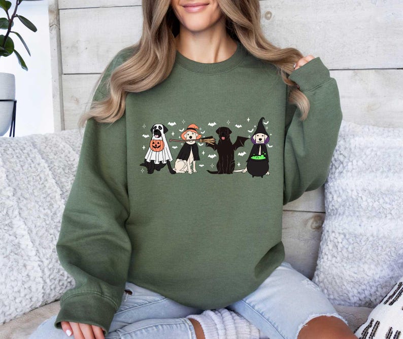 May include: A green sweatshirt with a graphic of four dogs dressed in Halloween costumes. The dogs are wearing a ghost costume, a witch costume, a vampire costume, and a pumpkin costume. The text "Happy Howl-oween" is printed on the sweatshirt.