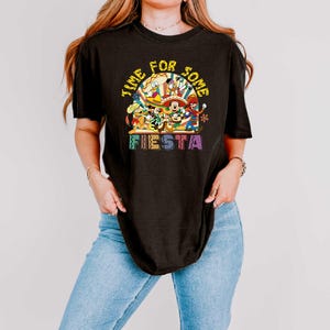 Pode incluir: Camiseta preta com um gr&aacute;fico colorido com personagens da Disney e o texto "Time for Some Fiesta". O design inclui Mickey Mouse, Pato Donald e outros personagens em trajes festivos. A camisa tem gola redonda e mangas curtas.
