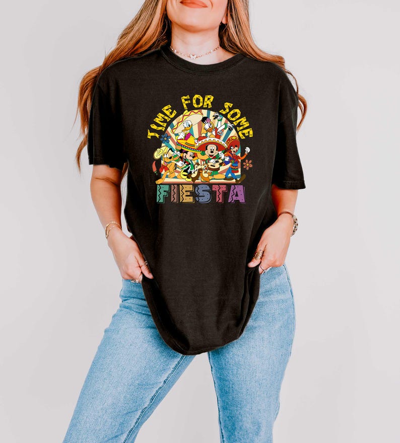 Pode incluir: Camiseta preta com um gr&aacute;fico colorido com personagens da Disney e o texto "Time for Some Fiesta". O design inclui Mickey Mouse, Pato Donald e outros personagens em trajes festivos. A camisa tem gola redonda e mangas curtas.