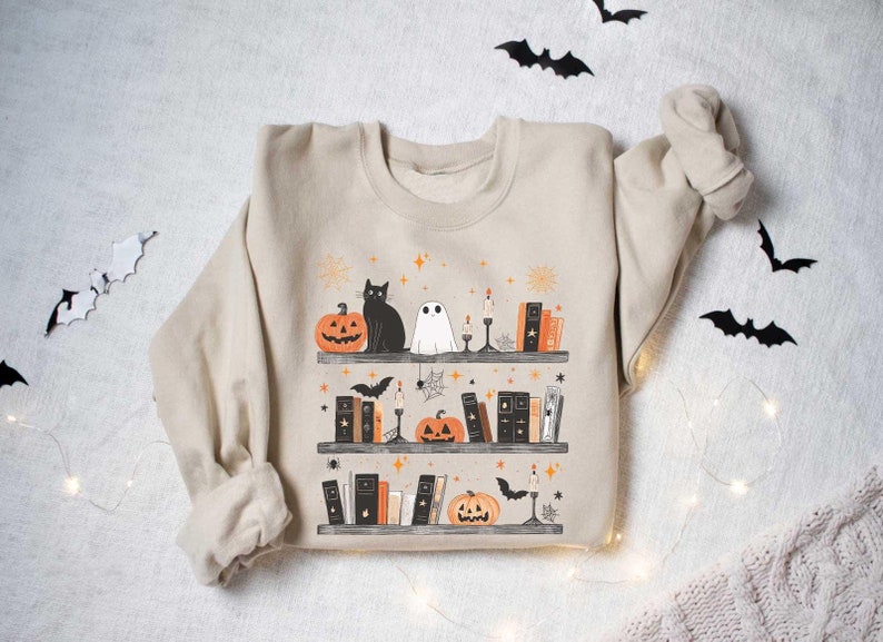 May include: A beige sweatshirt with a Halloween-themed design featuring a black cat, a ghost, pumpkins, and books on a shelf. The design is surrounded by stars and bats.