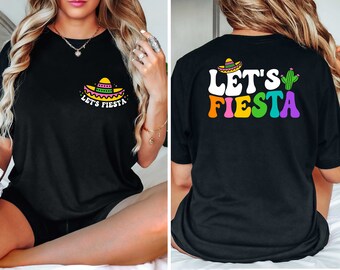 May include: Black t-shirt with a colorful "Let's Fiesta" design. The front features a sombrero graphic and text, while the back displays the phrase with a cactus illustration. The design incorporates vibrant colors, perfect for a festive occasion.