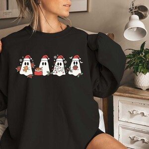 Op de afbeelding: Zwart sweatshirt met een afbeelding van vijf geesten die een kerstmanmuts dragen. De geesten houden geschenken, snoepstokken en een peperkoekman vast.