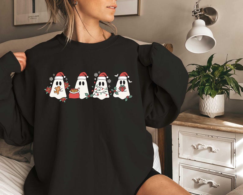 Op de afbeelding: Zwart sweatshirt met een afbeelding van vijf geesten die een kerstmanmuts dragen. De geesten houden geschenken, snoepstokken en een peperkoekman vast.