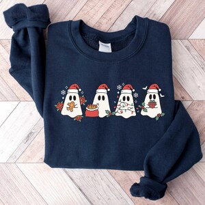 Op de afbeelding: Een donkerblauw sweatshirt met een kerstthema met vier geesten die een kerstmanmuts dragen. De geesten houden geschenken, snoepstokken en lampjes vast.