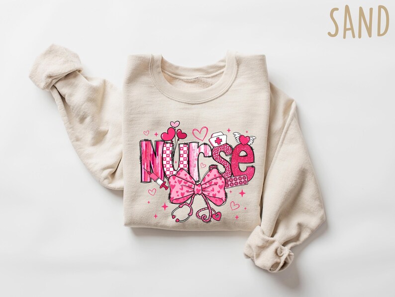 Peut inclure: Un sweat-shirt beige avec un motif graphique rose et blanc qui dit "Nurse" avec des c&oelig;urs, un st&eacute;thoscope et un bandage. Le sweat-shirt est pli&eacute; en deux.