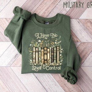 K&ouml;nnte beinhalten: Ein gr&uuml;ner Pullover mit dem Text "I Have No Shelf Control" und einer Grafik eines B&uuml;cherstapels mit Blumen.