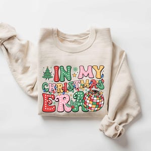 Op de afbeelding: Een beige sweatshirt met een kleurrijke grafische print die "In My Christmas Era" zegt met een discobal en kerstverlichting.