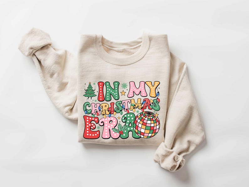 Op de afbeelding: Een beige sweatshirt met een kleurrijke grafische print die "In My Christmas Era" zegt met een discobal en kerstverlichting.