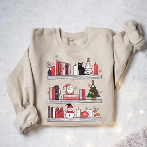 May include: A beige sweatshirt with a Christmas-themed design featuring a bookshelf with books, candles, a cat, a snowman, a Santa Claus, and a Christmas tree.