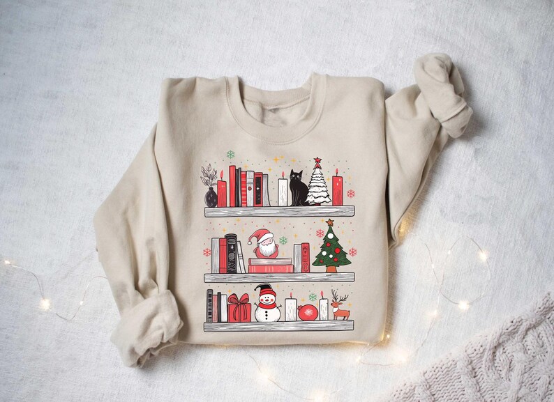 May include: A beige sweatshirt with a Christmas-themed design featuring a bookshelf with books, candles, a cat, a snowman, a Santa Claus, and a Christmas tree.
