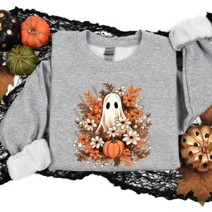 May include: A gray sweatshirt with a white ghost surrounded by orange, brown, and white flowers. The ghost is smiling and has a small pumpkin in its hand.