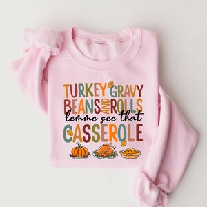 May include: Pink sweatshirt with a Thanksgiving-themed graphic that says "TURKEY GRAVY BEANS AND ROLLS lemme see that CASSEROLE". The graphic features a turkey, pumpkin, and pie.