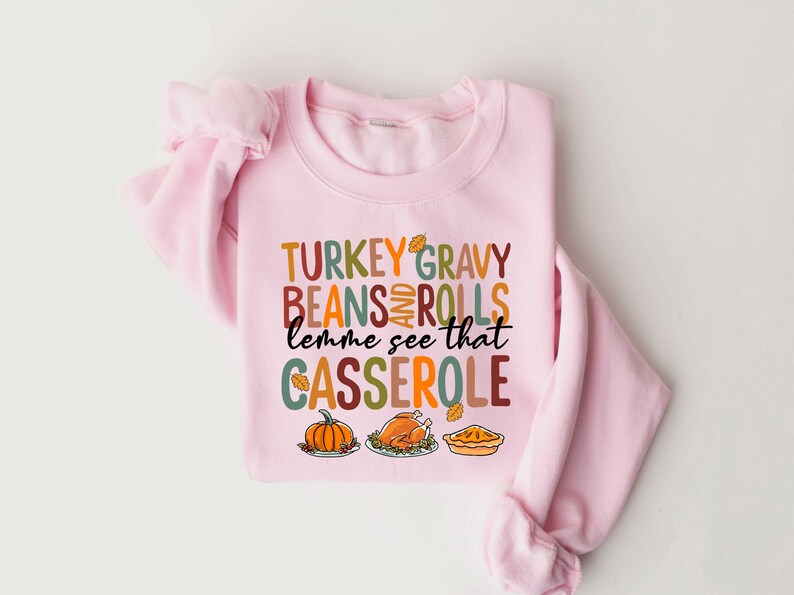 May include: Pink sweatshirt with a Thanksgiving-themed graphic that says "TURKEY GRAVY BEANS AND ROLLS lemme see that CASSEROLE". The graphic features a turkey, pumpkin, and pie.