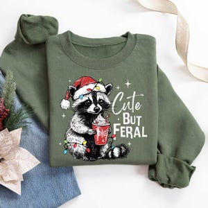 May include: Green Christmas sweatshirt with a cute raccoon wearing a Santa hat and holding a cup of hot chocolate. The text on the sweatshirt reads "Cute But Feral".