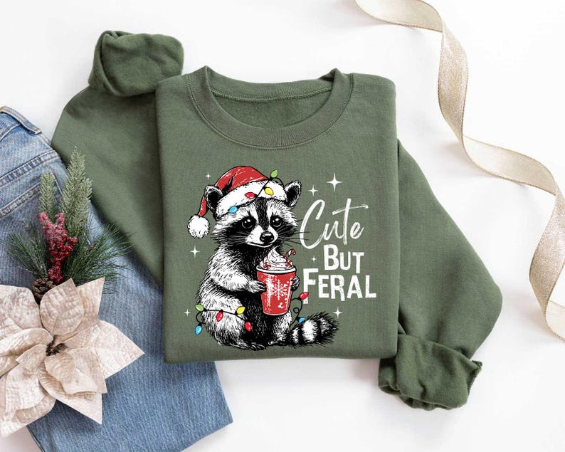 May include: Green Christmas sweatshirt with a cute raccoon wearing a Santa hat and holding a cup of hot chocolate. The text on the sweatshirt reads "Cute But Feral".