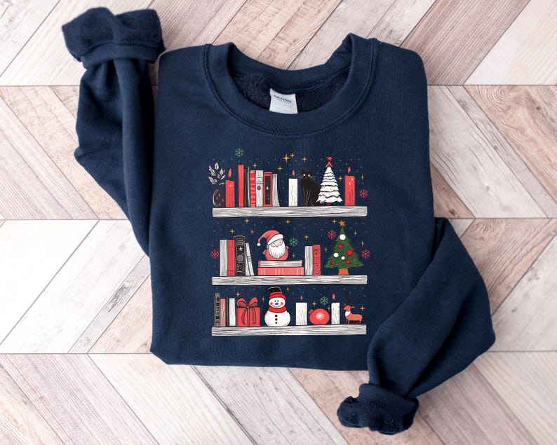 May include: A navy blue sweatshirt with a Christmas-themed design featuring a bookshelf filled with books, a snowman, a Santa Claus, and a Christmas tree. The design is in a white, red, and green colour scheme.