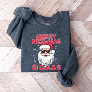 May include: A dark gray sweatshirt with the text "MERRY RIZZ-MAS SIGMAS" in red. The design features Santa Claus wearing sunglasses and a red hat. The sweatshirt is folded on a wooden surface.