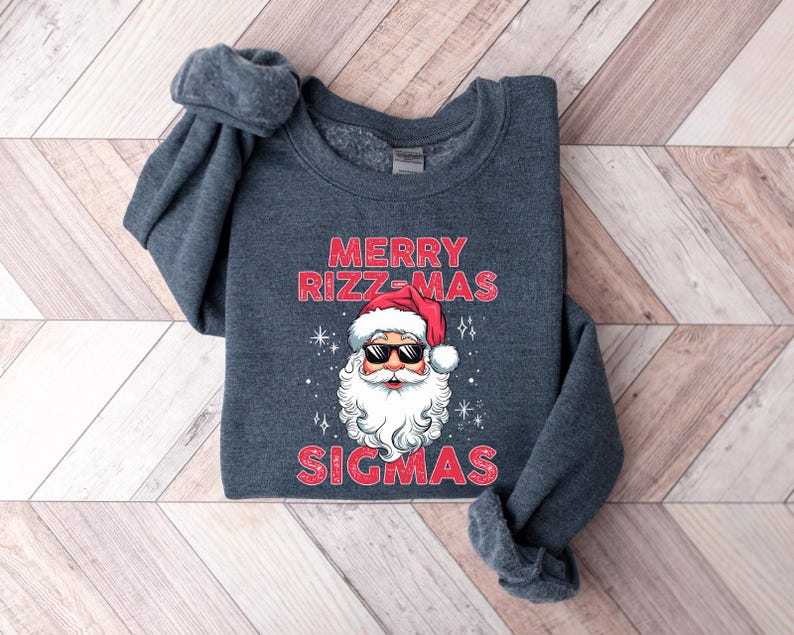 May include: A dark gray sweatshirt with the text "MERRY RIZZ-MAS SIGMAS" in red. The design features Santa Claus wearing sunglasses and a red hat. The sweatshirt is folded on a wooden surface.