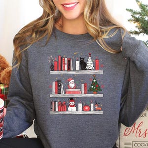 May include: A grey sweatshirt featuring a graphic of a bookshelf filled with books, Christmas decorations, a cat, Santa Claus, a snowman, and a Christmas tree. The text "Mrs. Claus Cookie Company" is printed on the bottom of the sweatshirt.