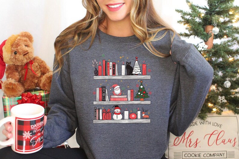 May include: A grey sweatshirt featuring a graphic of a bookshelf filled with books, Christmas decorations, a cat, Santa Claus, a snowman, and a Christmas tree. The text "Mrs. Claus Cookie Company" is printed on the bottom of the sweatshirt.