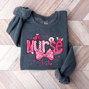 Peut inclure: Un sweat-shirt gris fonc&eacute; avec un motif graphique rose et blanc qui dit "Nurse" avec des c&oelig;urs, un st&eacute;thoscope et une seringue.