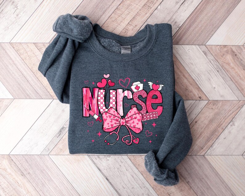 Peut inclure: Un sweat-shirt gris fonc&eacute; avec un motif graphique rose et blanc qui dit "Nurse" avec des c&oelig;urs, un st&eacute;thoscope et une seringue.