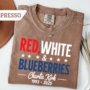 May include: Espresso-colored t-shirt with the words "RED, WHITE & BLUEBERRIES" in red, white, and blue. The shirt also includes the name "Charlie Kirk" and the years "1993-2025". A silver necklace is draped across the shirt.