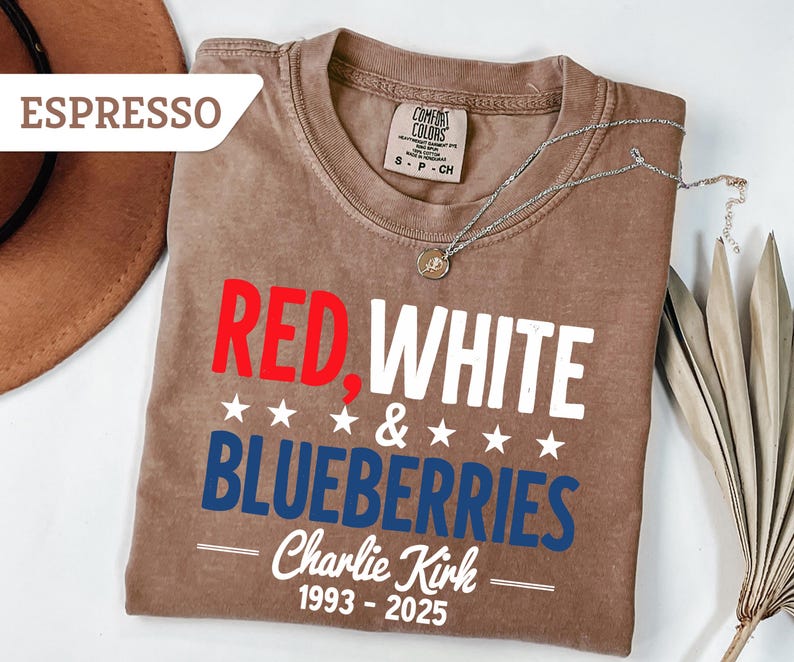 May include: Espresso-colored t-shirt with the words "RED, WHITE & BLUEBERRIES" in red, white, and blue. The shirt also includes the name "Charlie Kirk" and the years "1993-2025". A silver necklace is draped across the shirt.