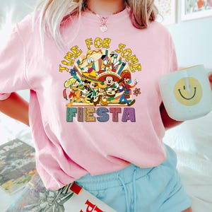 Pode incluir: Camiseta rosa claro com um gr&aacute;fico colorido apresentando personagens de desenhos animados e o texto "TIME FOR SOME FIESTA". O design inclui Mickey Mouse, Pato Donald e outros personagens em trajes festivos.
