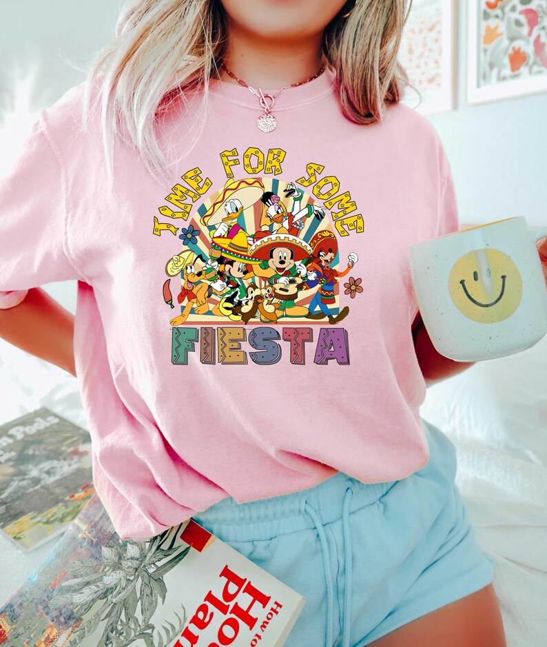 Pode incluir: Camiseta rosa claro com um gr&aacute;fico colorido apresentando personagens de desenhos animados e o texto "TIME FOR SOME FIESTA". O design inclui Mickey Mouse, Pato Donald e outros personagens em trajes festivos.