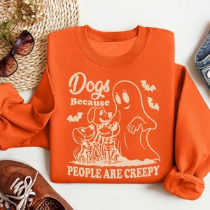 Puede incluir: Sudadera naranja con un estampado gr&aacute;fico de un fantasma y tres perros. El texto en la sudadera dice "Dogs Because People Are Creepy".