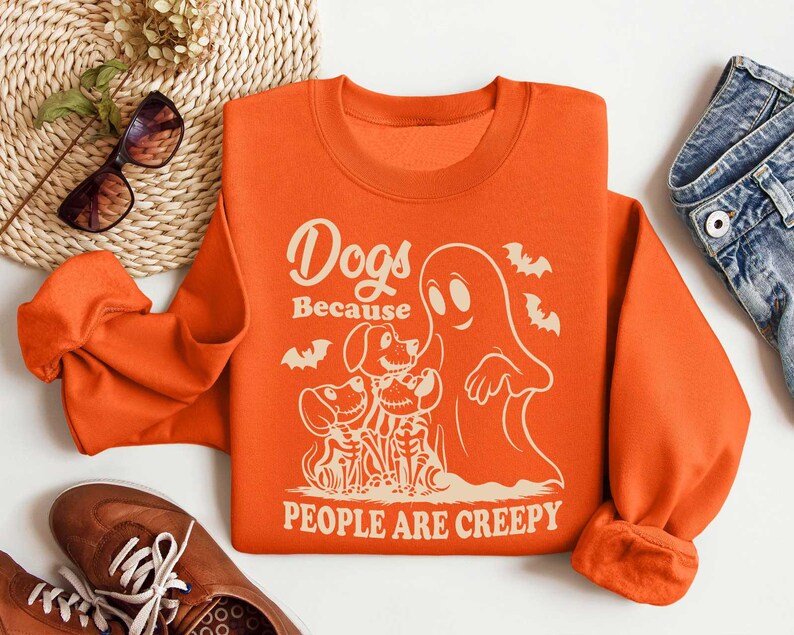 Puede incluir: Sudadera naranja con un estampado gr&aacute;fico de un fantasma y tres perros. El texto en la sudadera dice "Dogs Because People Are Creepy".