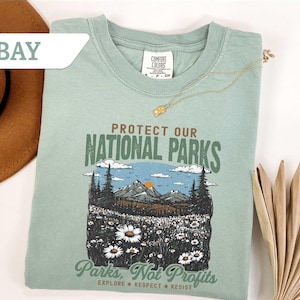 Może przedstawiać: Jasnozielona koszulka z napisem "PROTECT OUR NATIONAL PARKS" i grafiką górskiego krajobrazu z drzewami i kwiatami. Na koszulce wydrukowano również tekst "Parks, Not Profits". Złoty naszyjnik jest ułożony na koszulce.