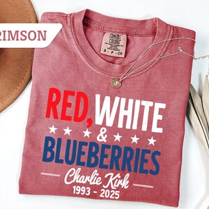 May include: Crimson-colored t-shirt with the text "RED, WHITE & BLUEBERRIES" in red, white, and blue. The shirt also includes the name "Charlie Kirk" and the years "1993-2025". A silver necklace with a pendant is draped across the shirt.