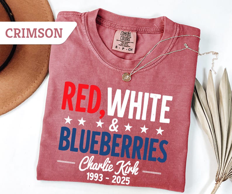 May include: Crimson-colored t-shirt with the text "RED, WHITE & BLUEBERRIES" in red, white, and blue. The shirt also includes the name "Charlie Kirk" and the years "1993-2025". A silver necklace with a pendant is draped across the shirt.