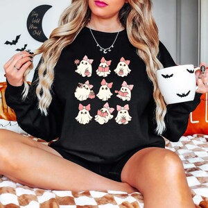 May include: Black sweatshirt featuring a repeating pattern of cartoon ghosts with pink bows. The ghosts are holding books, flowers, and gifts. A white mug with bat silhouettes is held in the hand.