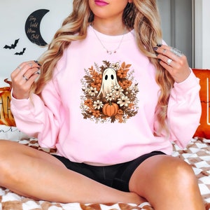 May include: A pink sweatshirt with a white ghost holding a small orange pumpkin surrounded by fall flowers. The ghost is in the center of the design and the flowers are in a circle around it.