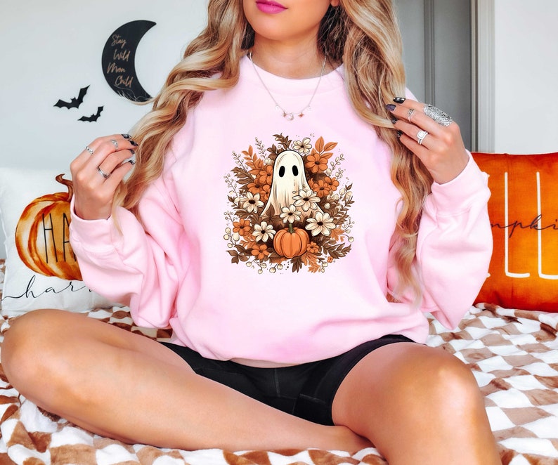 May include: A pink sweatshirt with a white ghost holding a small orange pumpkin surrounded by fall flowers. The ghost is in the center of the design and the flowers are in a circle around it.