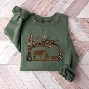 Puede incluir: Una sudadera verde con un gr&aacute;fico marr&oacute;n que representa una escena de la Natividad, con una estrella, monta&ntilde;as y &aacute;rboles. El dise&ntilde;o incluye un caballo, una persona arrodillada y un beb&eacute; en un pesebre. Las mangas est&aacute;n dobladas.